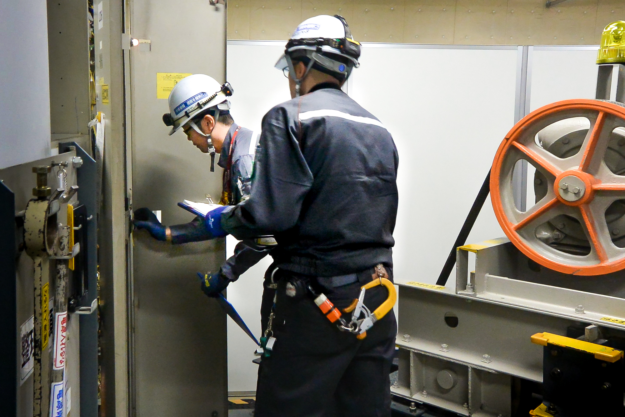 Mr. Umemura (at the background) attempting to open the control panel of a conventional type of elevator, which is located in Field Services Training Center, to measure the insulation resistance, during a legal inspection work competition in 2017.