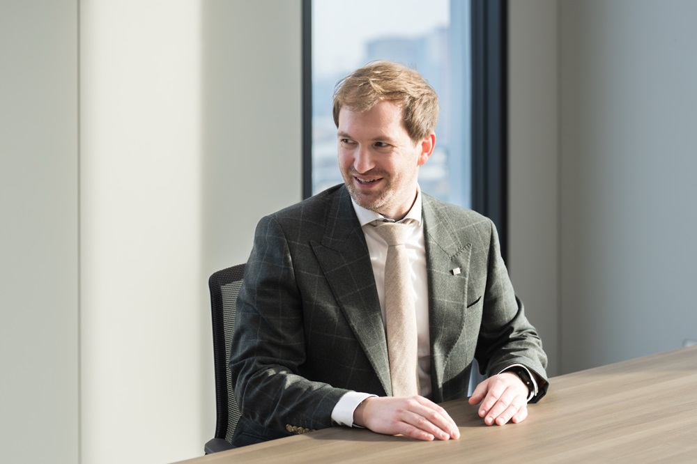 Dr. Thomas Roger seated at a table in a bright office, looking to the side with a slight smile during a conversation.