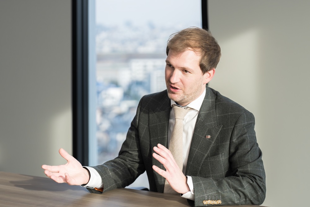  Dr. Thomas Roger engaged in conversation, seated at a table and gesturing with his hands in a bright office environment.