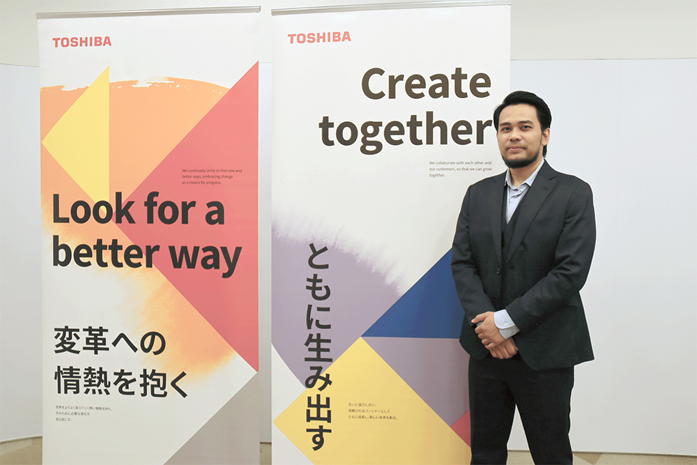 Angga Suherman posing next to Toshiba banners highlighting the company values “Look for a better way” and “Create together.”